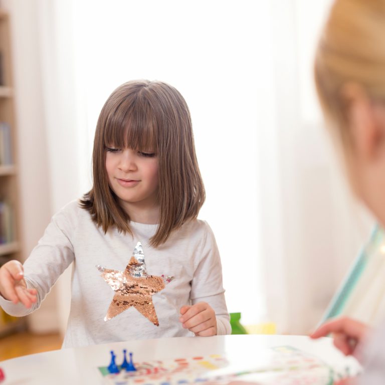 Mother and daughter sitting in a playroom, playing a ludo game; daughter rolling the dice